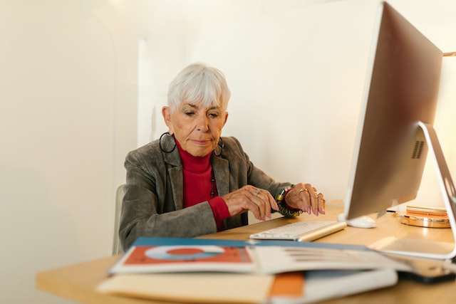 Frau sitzt am Schreibtisch vor einem Monitor. Sie betrachtet verschiedene Unterlagen die neben ihr liegen.