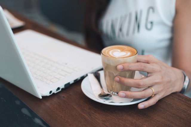 Eine Frau sitzt am Laptop beim Online-Netzwerken. Daneben steht ihr Kaffee den sie gerade in die Hand nimmt.
